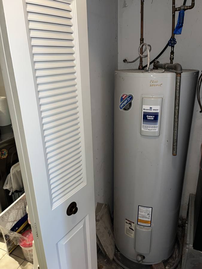 Old electric water heater in a utility closet with a rusted steel relief pipe